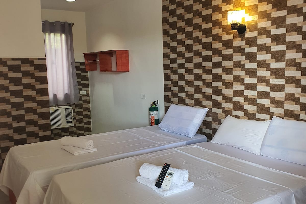 Una camera d'albergo con due letti, lenzuola bianche, un lampadario a parete e il banco della reception visibile attraverso una porta aperta.