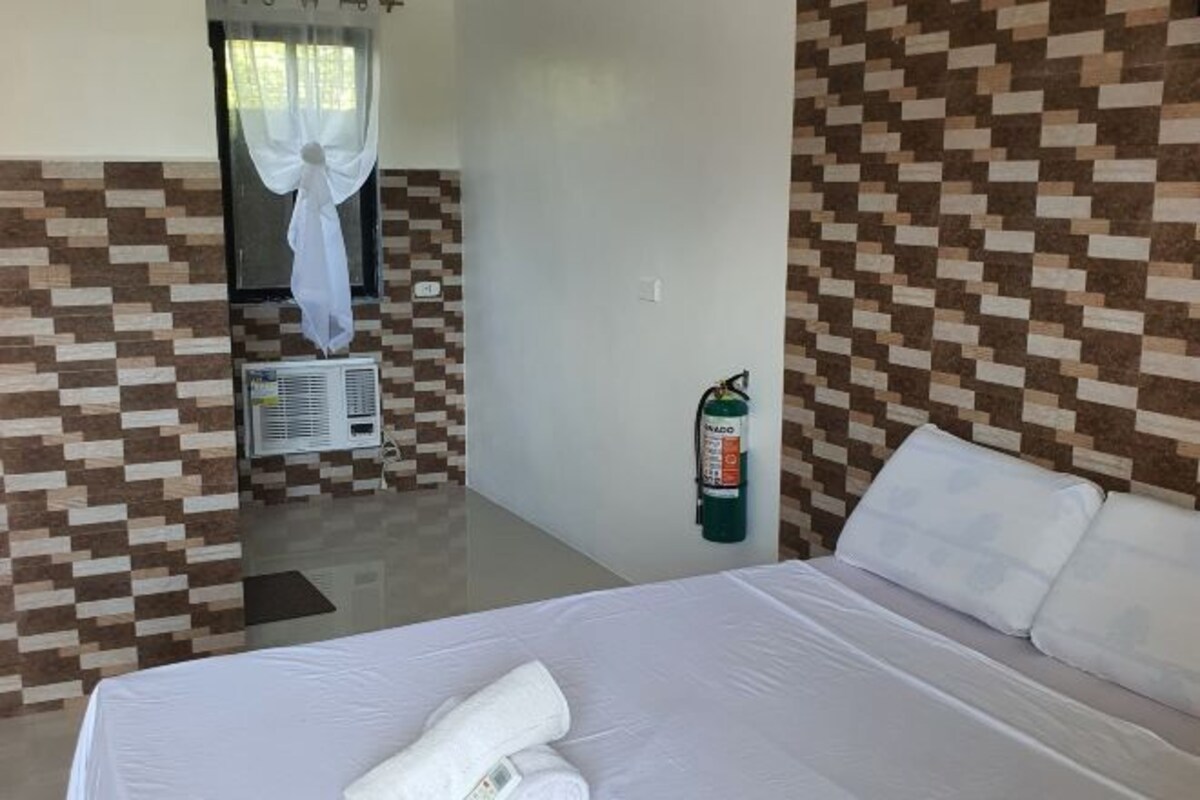 Ett hotellrum med en säng, en väggmonterad luftkonditioneringsanläggning, en brandsläckare och ett fönster med en tunn gardin.