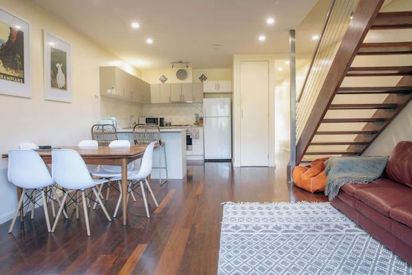 A modern kitchen with white cabinets, a dining area with a wooden table and chairs, a sofa with a throw blanket, and a staircase.