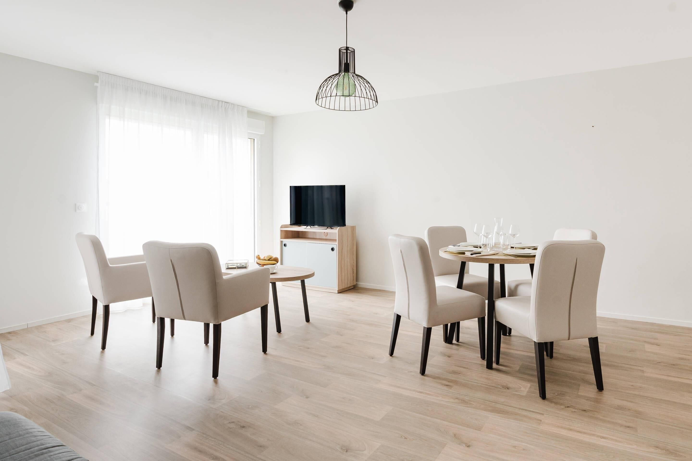 Een moderne eetruimte met een ronde tafel, zes stoelen, een televisie en een groot raam met dunne gordijnen.