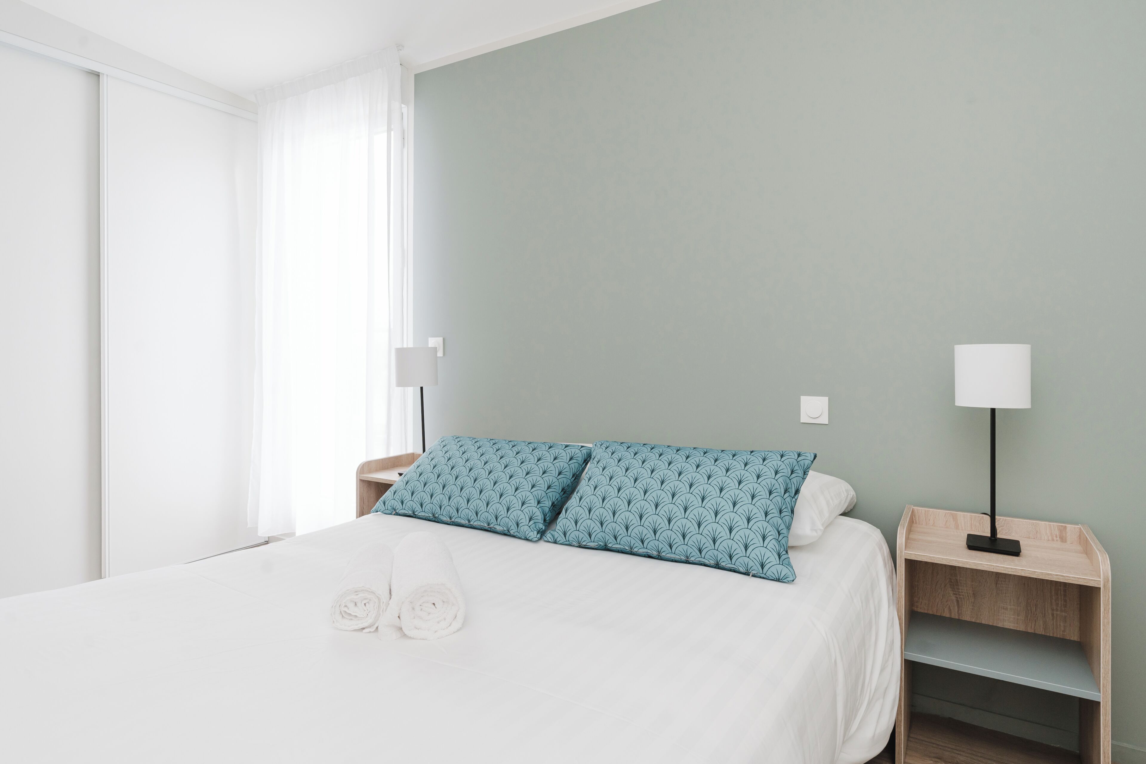 A neatly made bed with two pillows, a bedside table with a lamp, and a window with curtains.