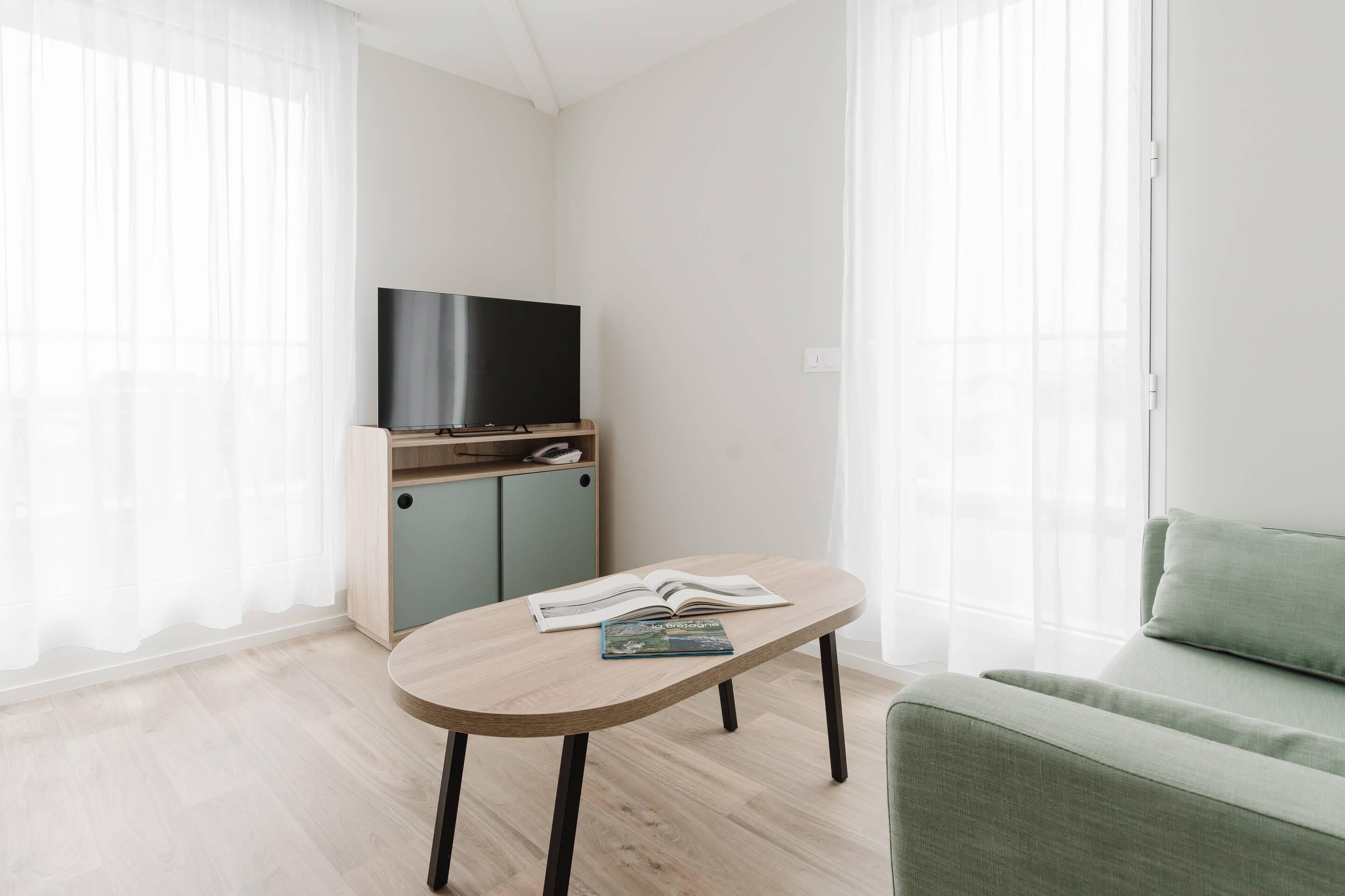 Un salon moderne avec un téléviseur à écran plat posé sur un meuble en bois, un canapé de couleur claire et une petite table sur laquelle se trouve un livre.