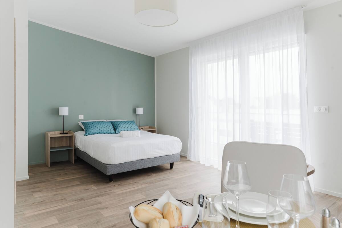 Ein modernes Hotelzimmer mit einem Bett, Nachttischen, einem Esstisch mit Tellern und Gläsern sowie einem Fenster mit leichtem Vorhang.