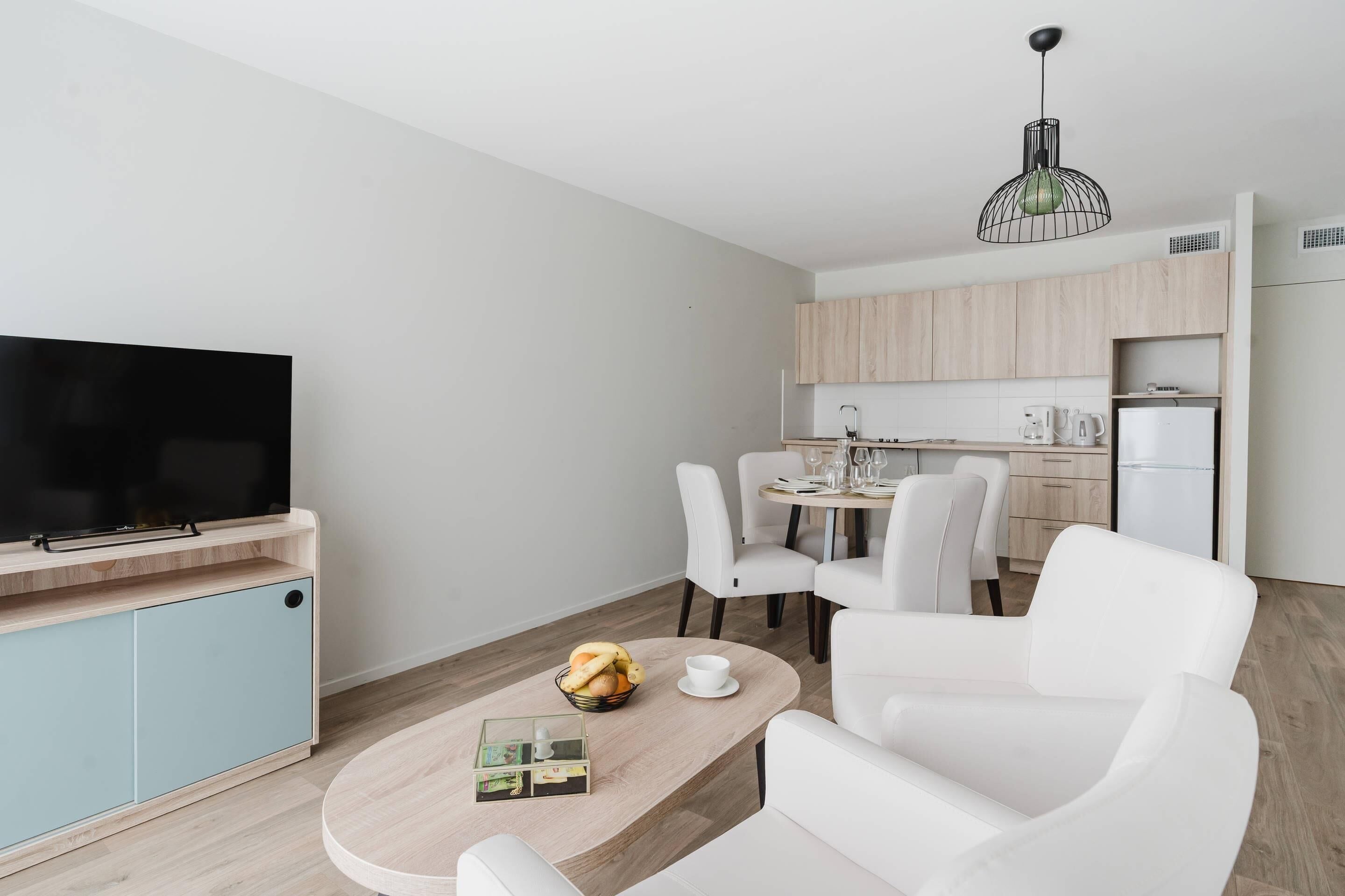 A modern living room with a flat-screen TV, a wooden coffee table, and a dining area with white chairs and a kitchen in the background.