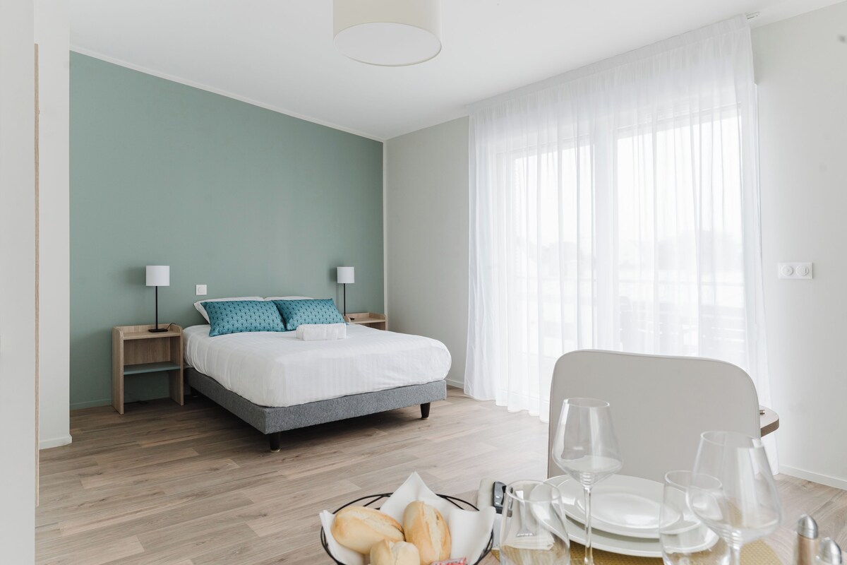 A modern hotel room with a bed, bedside tables, a dining table with plates and glasses, and a window with sheer curtains.