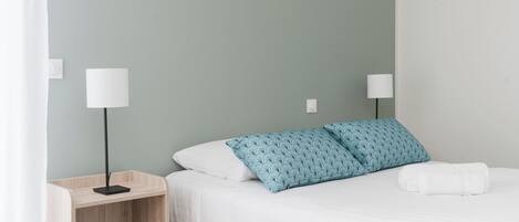 Ein ordentlich bezogenes Bett mit weißer Bettwäsche und einem blau gemusterten Kissen, ein Nachttisch mit Lampe sowie ein an der Wand befestigter Lichtschalter.