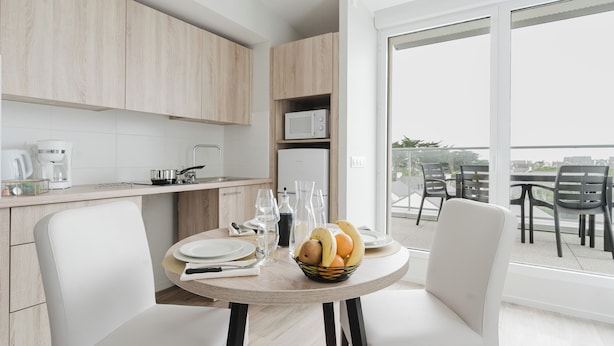 Une cuisine moderne avec des meubles en bois, une table à manger agrémentée d’un panier de fruits et un balcon avec un coin salon extérieur.