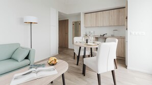 Un salon moderne avec un canapé, une table à manger et des chaises, une table basse avec des magazines, et un coin cuisine à l’arrière-plan.
