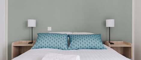 Una cama bien hecha con sábanas blancas y una toalla blanca enrollada, flanqueada por dos mesitas de noche con lámparas y un cojín estampado.
