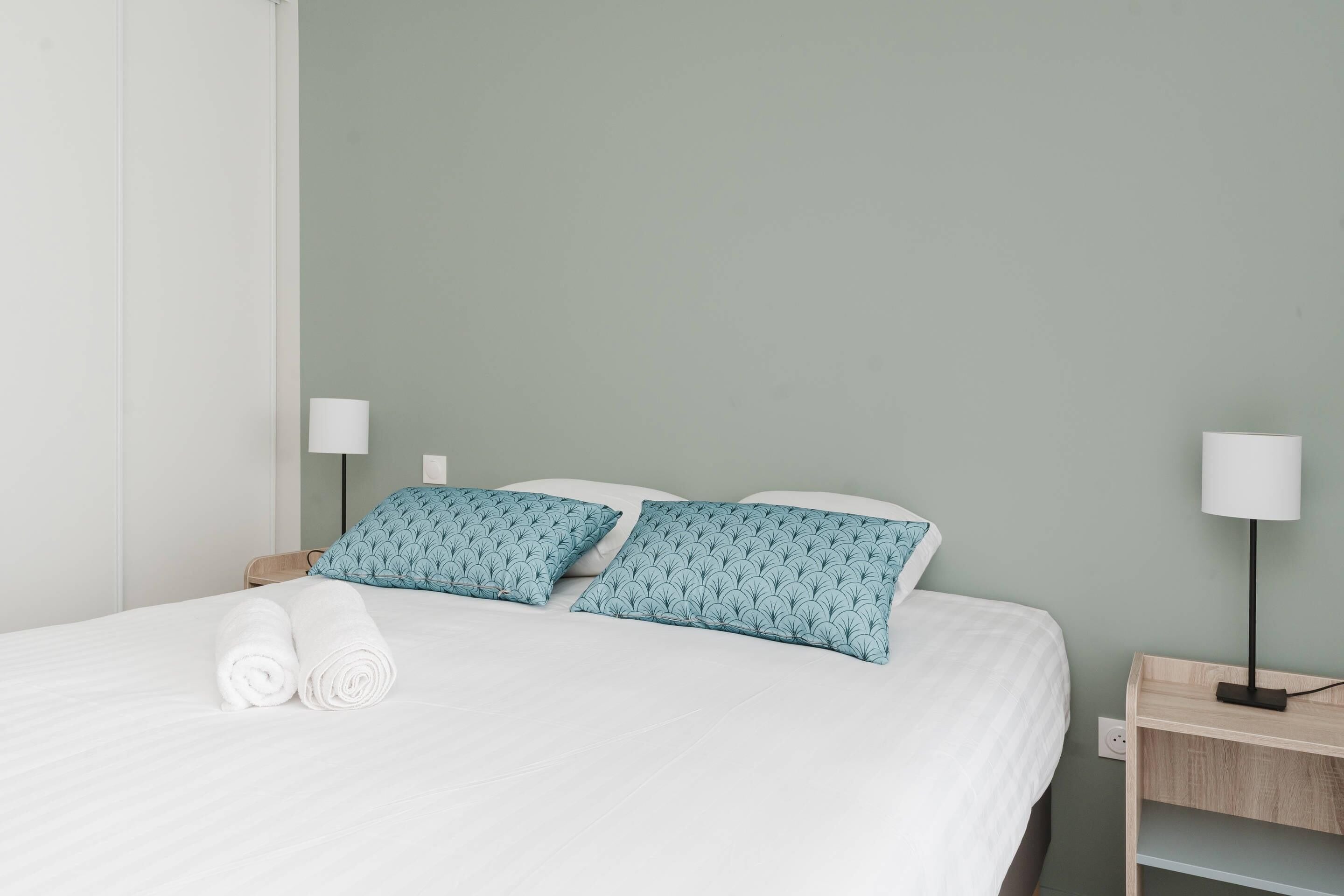 A neatly made bed with white bedding and two teal pillows, a bedside table with a lamp, and a wall socket.