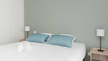 A neatly made bed with white bedding and two teal pillows, a bedside table with a lamp, and a wall socket.