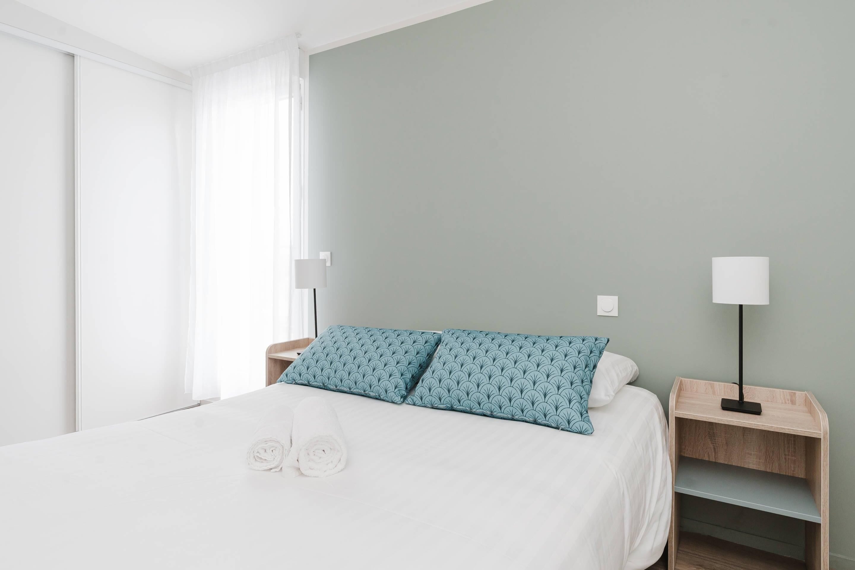 Un lit bien fait, avec une literie blanche et deux coussins à motifs bleus, une table de chevet avec une lampe, et une fenêtre ornée de rideaux légers.