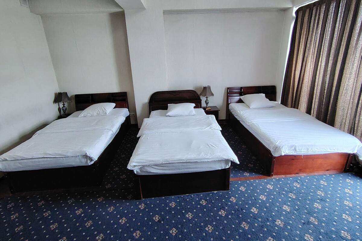Ein Hotelzimmer mit drei Einzelbetten, einem blauen Teppich mit Muster und einem großen Fenster mit Vorhängen.