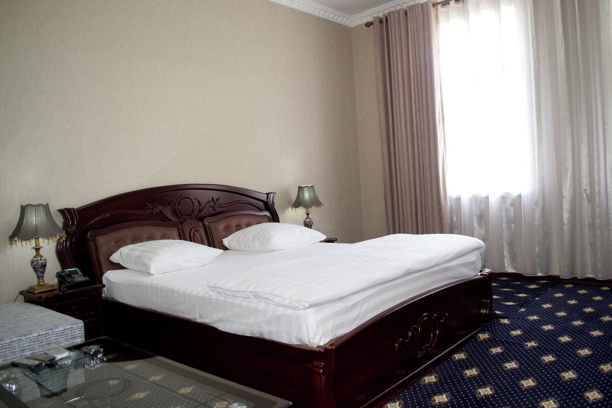 Ein Hotelzimmer mit einem großen Bett, zwei Nachttischen mit Lampen, einem Glastisch und einem Fenster mit Vorhängen.