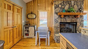 A wooden-paneled room with a stone fireplace, a wooden table with a chair, and a clock on the wall. - Cozy Cove Retreat by Chatuge Home Concierge | Private Swim Dock, Kayaks & Grill (Hayesville)