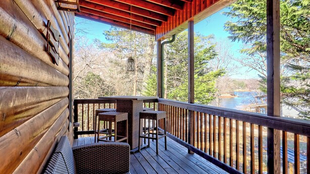 A wooden deck with wicker furniture and a view of a forest and water. - Cozy Cove Retreat by Chatuge Home Concierge | Private Swim Dock, Kayaks & Grill (Hayesville)