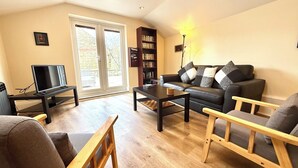 Un salon avec un canapé, une table basse et des fauteuils. On y trouve également un téléviseur posé sur un meuble, une bibliothèque et une lampe.