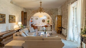 A cozy living room with a sofa, a coffee table, and a chandelier. There is a stone wall and a wooden cabinet. - Villa la Pizzichina 10 Emma Villas (Todi)