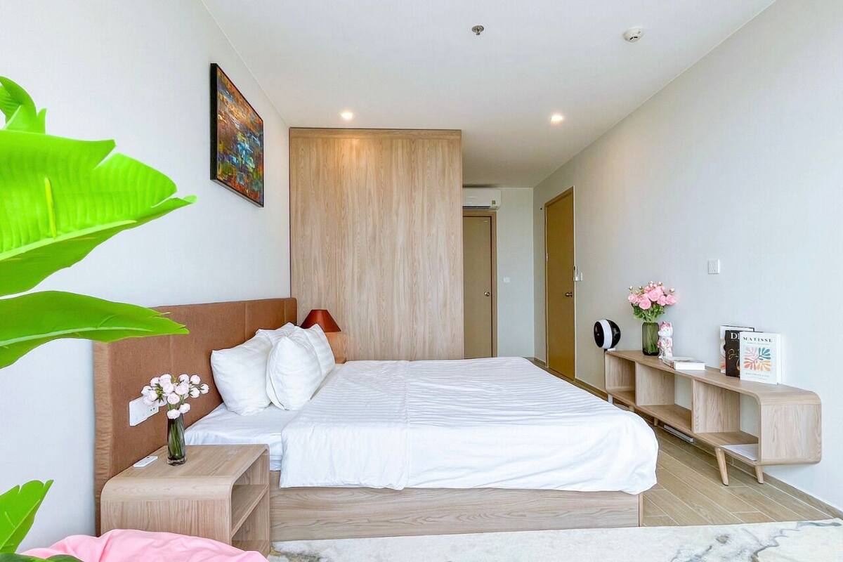 Une chambre moderne avec un grand lit, une tête de lit en bois, des tables de chevet, une petite table de nuit, une console agrémentée d’objets décoratifs et une grande plante verte.