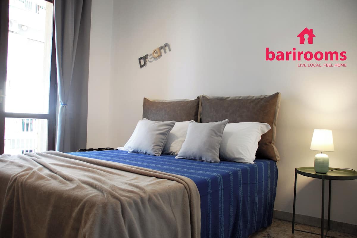 A neatly made bed with a blue and beige bedspread, a bedside lamp, and a wall with the words "Dream" and "barrirooms" logo.