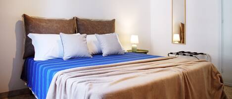 Un lit bien fait, recouvert d’une couvre-lit bleue et beige, avec deux oreillers marron et un oreiller blanc. Une table de chevet verte se trouve à côté du lit, et un miroir est accroché au mur.