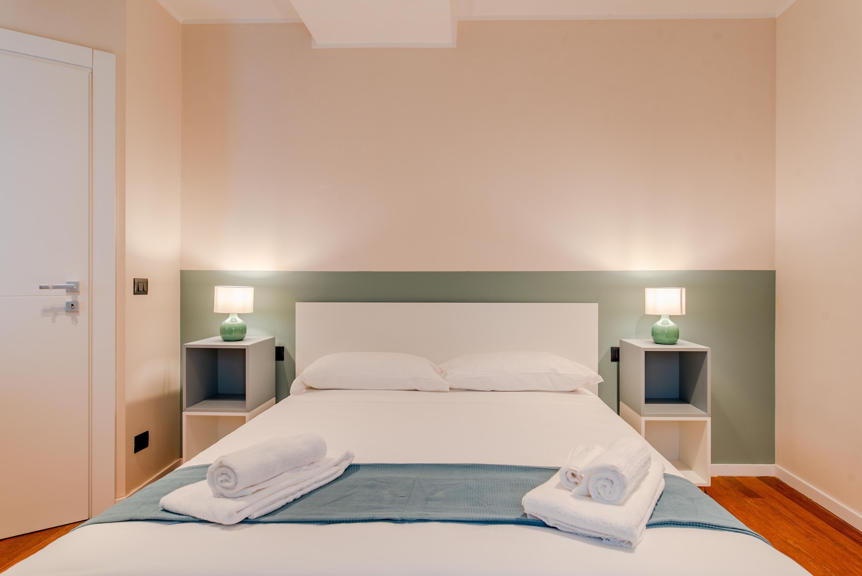 A neatly made bed with white linens, two bedside tables with lamps, and a door to the left.