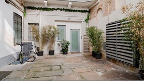 Une cour avec un sol en pierre, des plantes en pot, un vélo et une porte dotée d’un cadre vert.