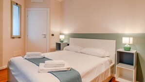 Un lit bien fait, avec du linge de lit blanc et une serviette pliée, une table de chevet avec une lampe, et une porte avec une fenêtre.
