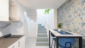 Una cocina moderna con encimera de mármol, armarios blancos y una escalera que lleva a un comedor con una silla azul y una planta.
