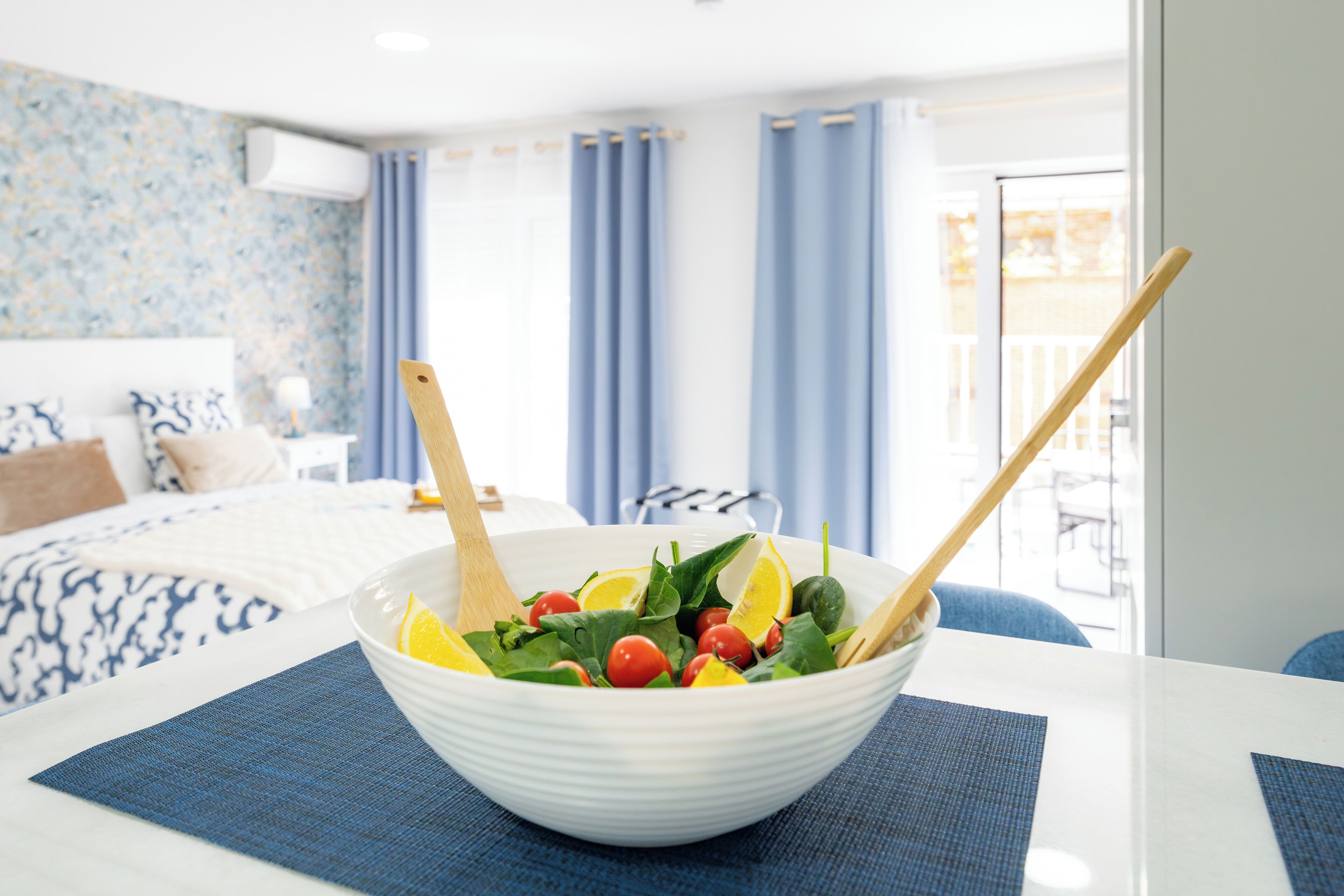 Un tazón blanco con ensalada sobre una mesa, una cama con ropa de cama estampada y un balcón con cortinas azules al fondo.