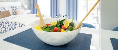 A white bowl with a salad on a table, a bed with patterned bedding, and a balcony with blue curtains in the background.