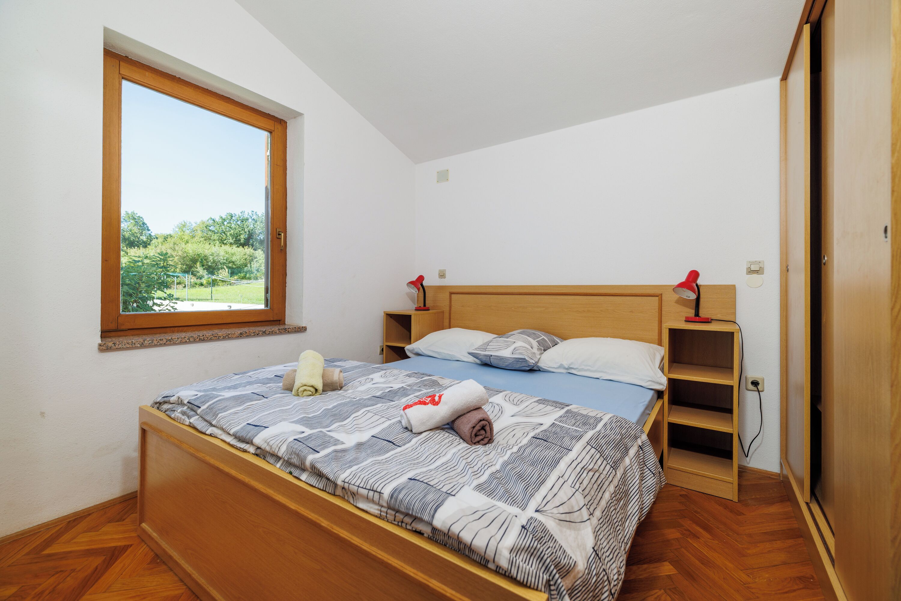 Une chambre avec un lit en bois, une fenêtre donnant sur un paysage verdoyant, une table de chevet avec une lampe et une étagère intégrée.
