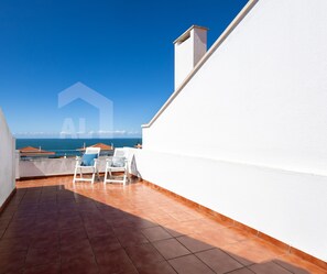 Un balcón con vista al mar, paredes blancas y dos sillas blancas.