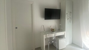 A modern, minimalist room with a white desk, a flat-screen TV mounted on the wall, and a window with sheer curtains. - Mamuccia Rooms (Naples)