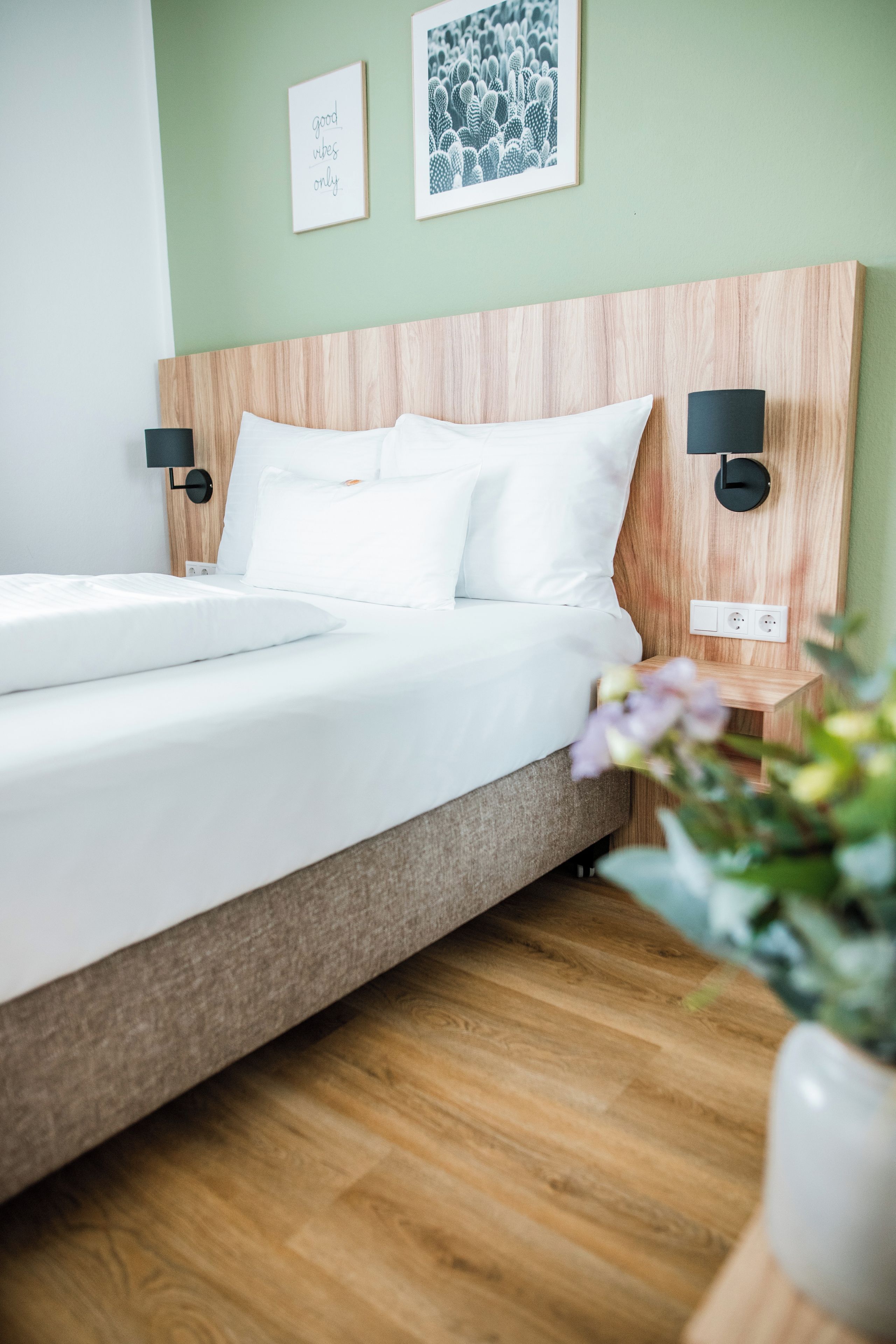 A modern hotel room with a wooden headboard, white bedding, two wall-mounted nightstands, and a potted plant.