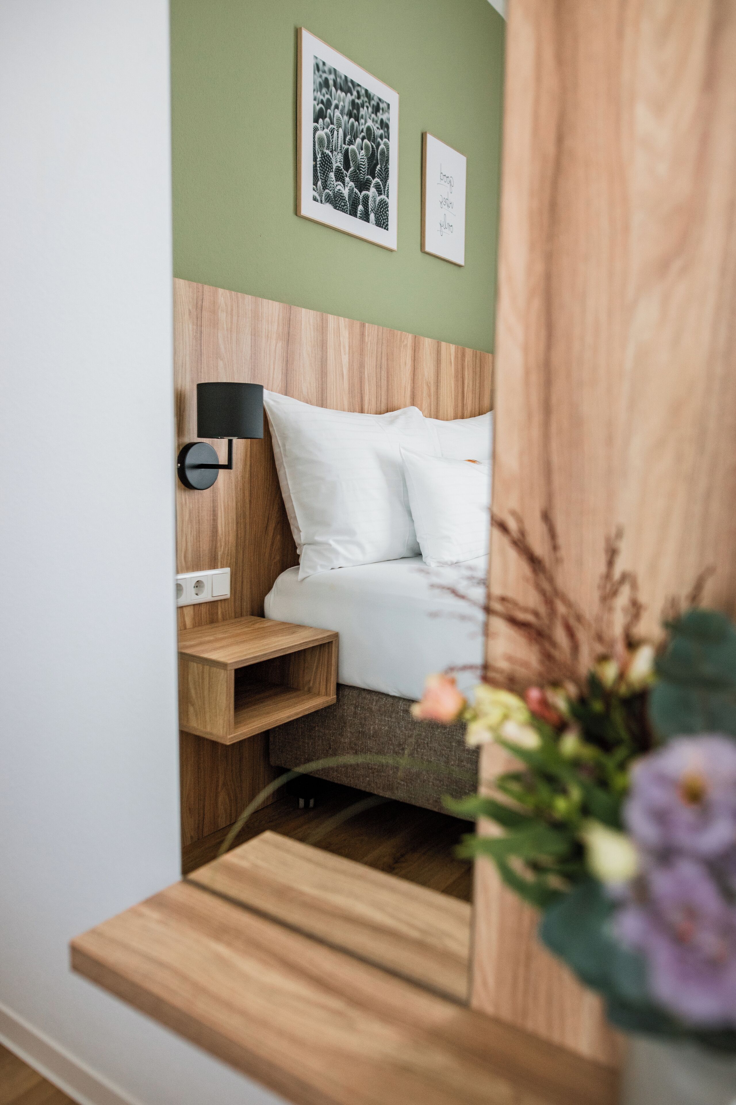 A bedroom with a wooden headboard, a white bed, a bedside table, a black lamp, and a small shelf.