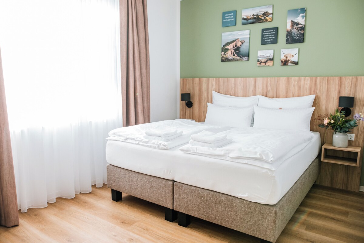 A neatly made bed with white linens and pillows, a wooden headboard, and a small bedside table with a vase of flowers.
