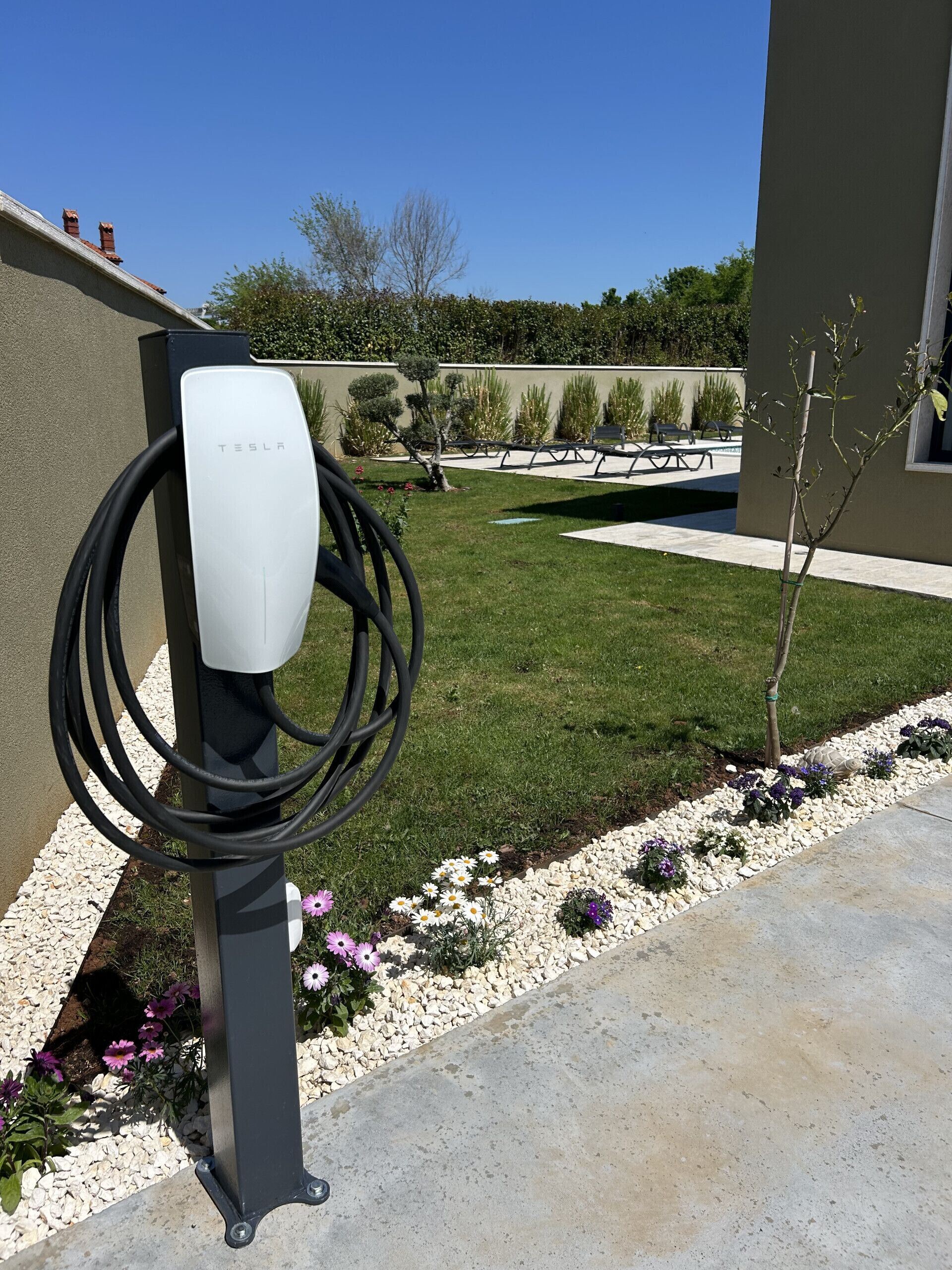 A Tesla charging station in a landscaped outdoor area with a patio, garden beds, and a building in the background.