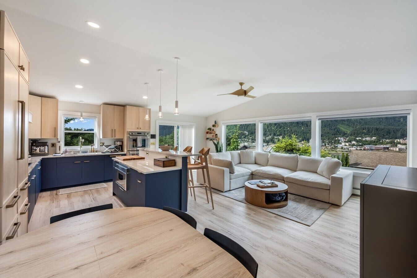 A modern kitchen with a central island, a dining area with a round table, a living room with a sofa and a coffee table, and large windows with a view of the outdoors.