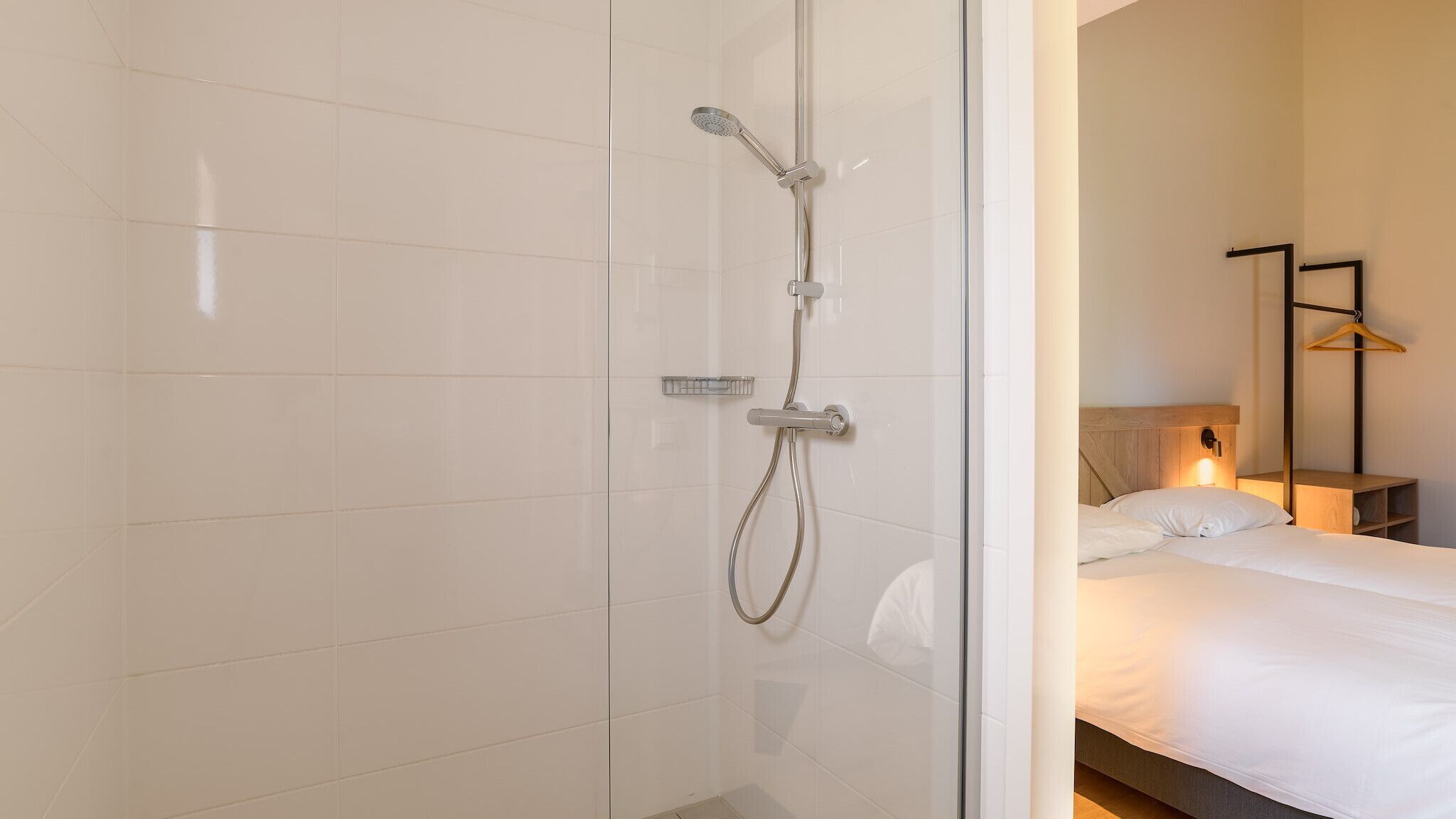 A modern bathroom with a glass shower enclosure, a bedside table with a lamp, and a neatly made bed.