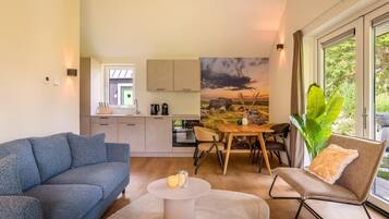 Un salon moderne avec un canapé, une table basse et un coin repas comprenant une table et des chaises. Au fond, on aperçoit une cuisine avec des placards et une fenêtre.