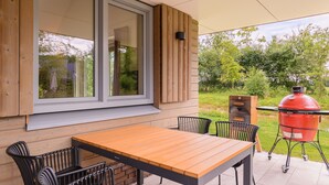 Een terras buiten met een houten tafel en stoelen, een groot raam en een rode barbecue.