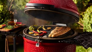 Un barbecue rouge, portable, avec le couvercle ouvert, sur lequel on fait griller des légumes et un morceau de viande.
