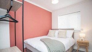 A bedroom with a bed, a bedside table with a lamp, a window with blinds, and a wall-mounted coat rack.