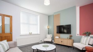 A modern living room with a sofa, a round coffee table, a TV mounted on the wall, and a window with blinds.