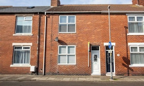 A red brick residential building with a white door, windows with shutters, and a blue street sign. - Meldon Terrace | Parking | Wi-Fi (Northumberland)