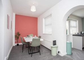 A dining area with a white table and chairs, a red accent wall, a radiator, a small trash bin, and a TV on a stand. - Meldon Terrace | Parking | Wi-Fi (Northumberland)