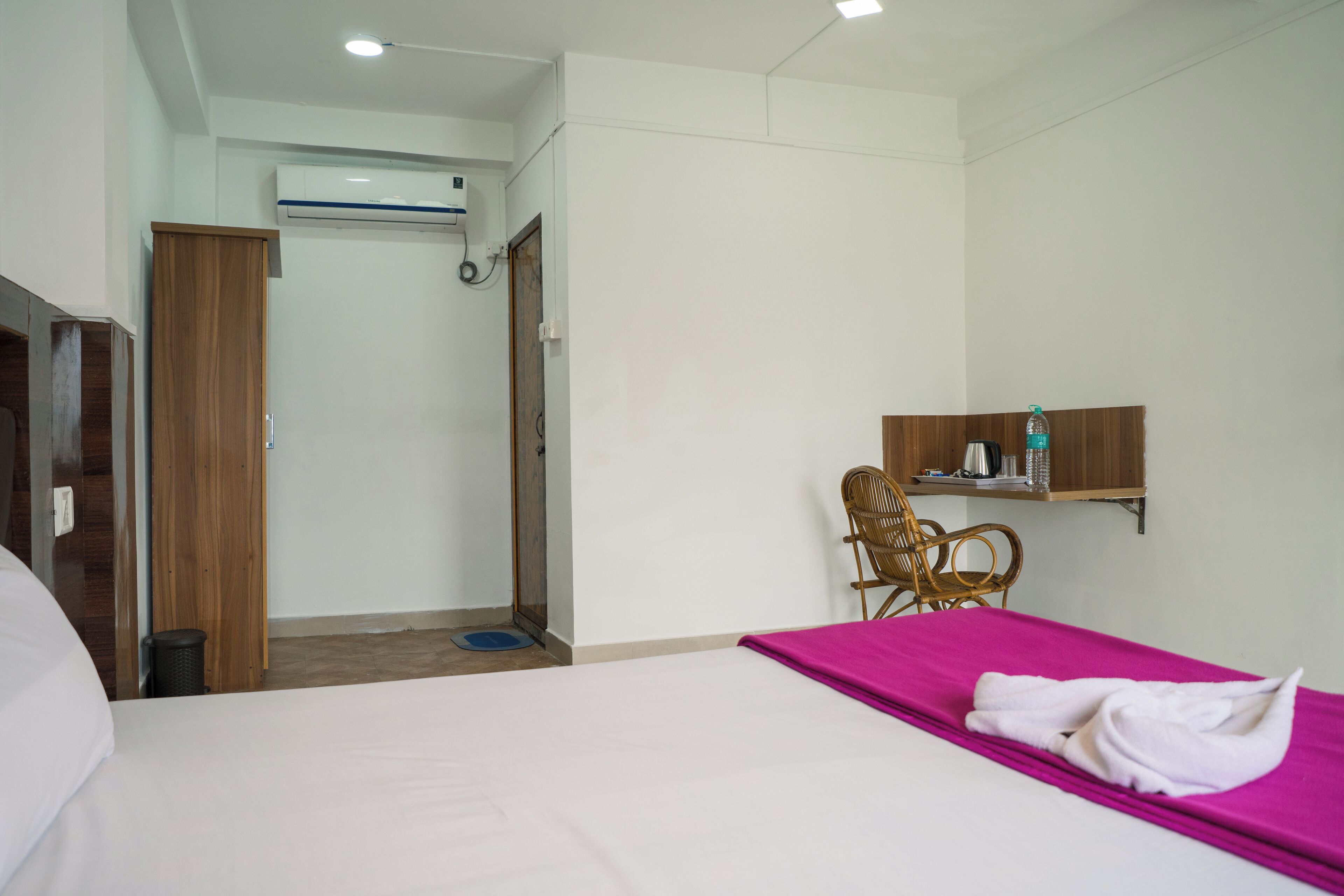A hotel room with a bed, a wooden wardrobe, a bathroom with a shower, and a small seating area with a chair.