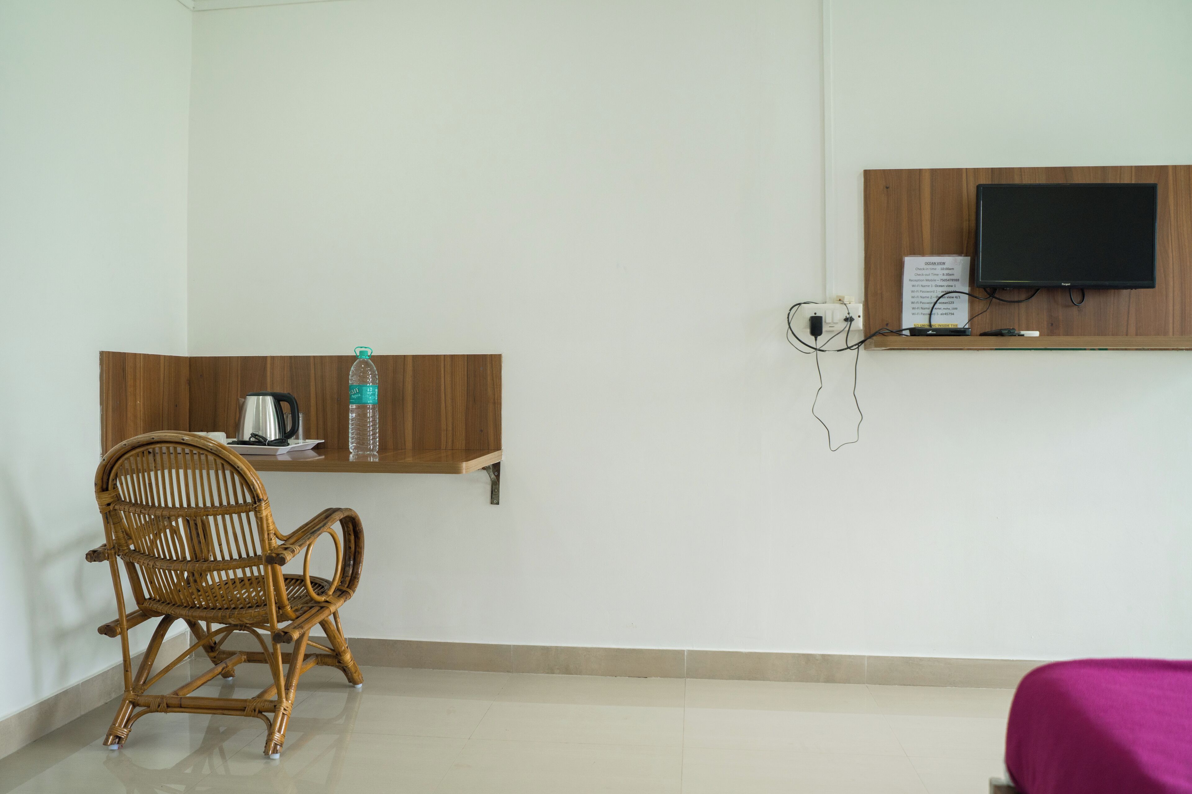 A room with a wicker chair, a wooden shelf with a water bottle, a kettle, and a flat-screen TV mounted on the wall.