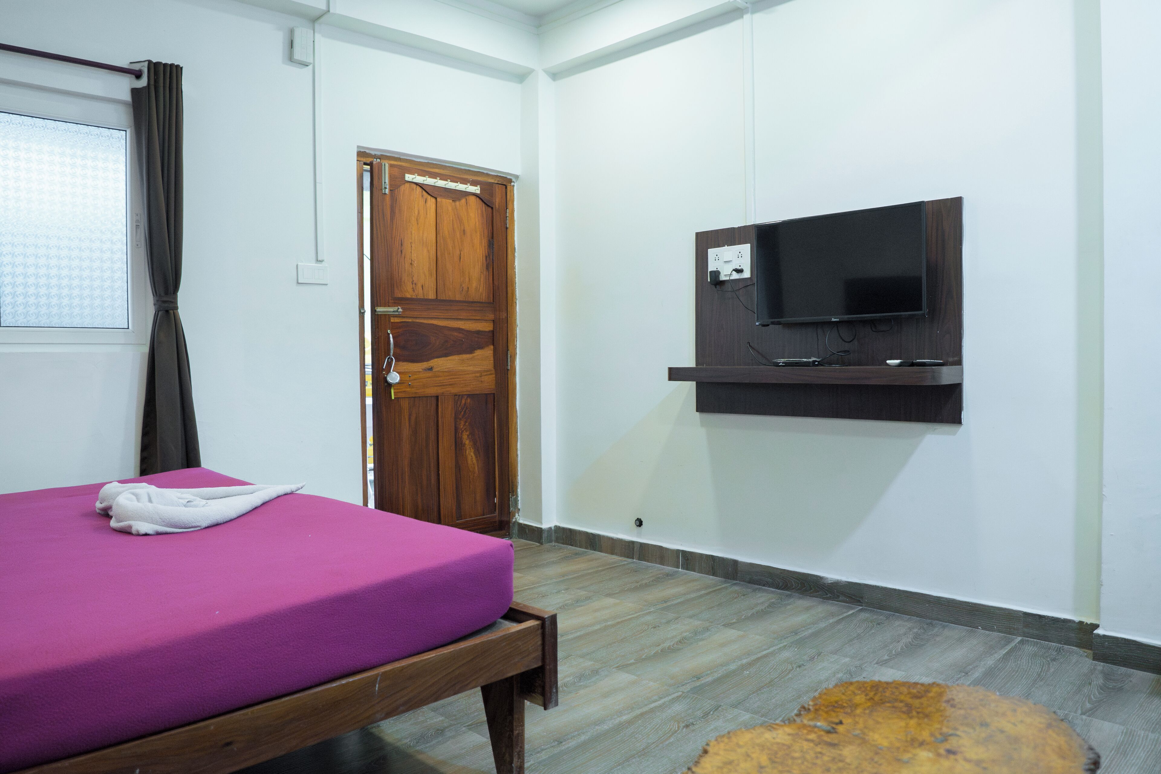 Une pièce avec un lit, une porte en bois, un téléviseur fixé au mur et une petite table ronde.